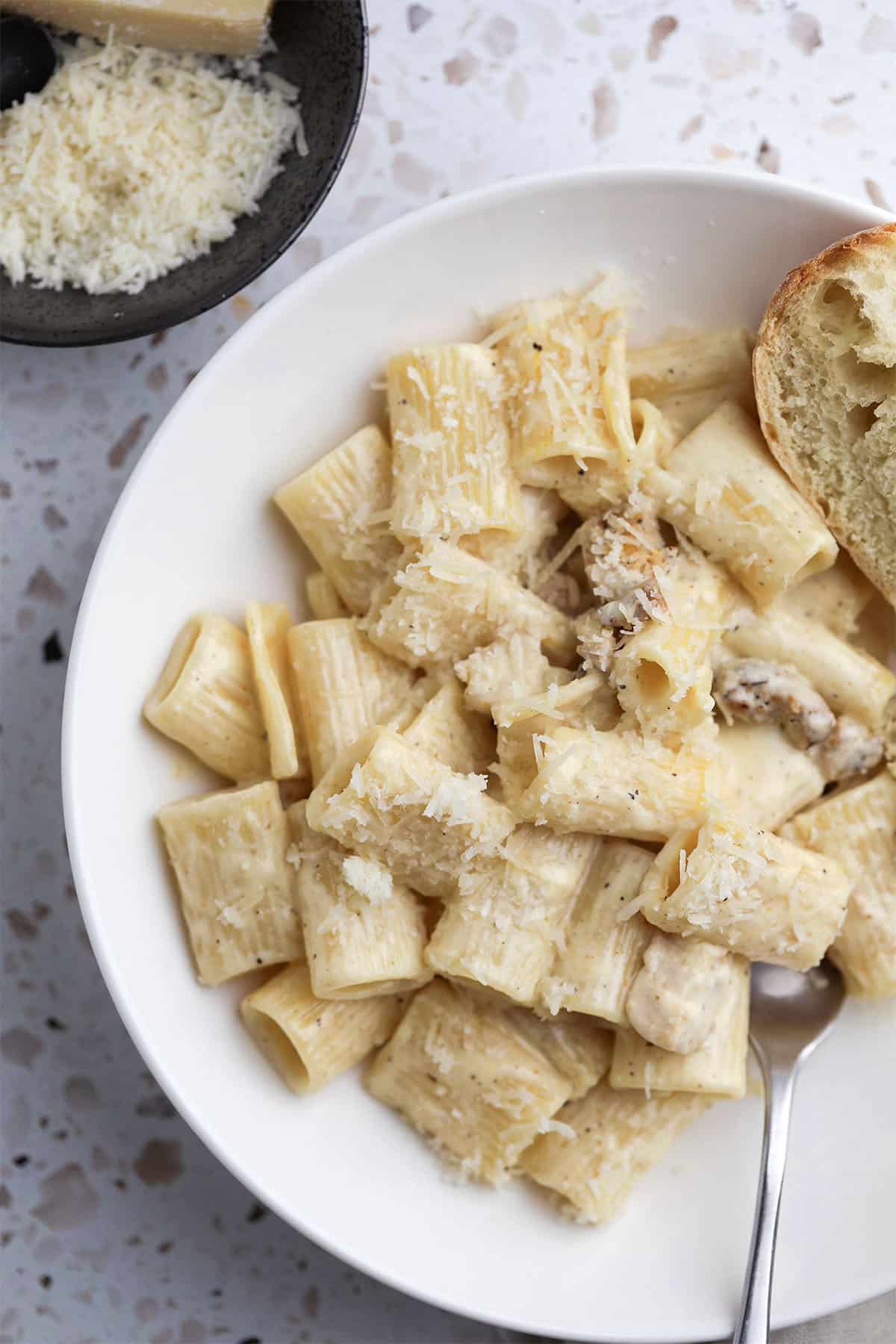 A white bowl of rigatoni pasta in a creamy homemade alfredo sauce with grated cheese and chicken, served with a slice of bread. A fork rests in the bowl, and a dish of grated cheese is visible in the background.