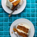 Two white plates with slices of carrot cake topped with white frosting and served with forks, placed on a bright blue tiled surface—perfect for showcasing the best carrot cake recipe.