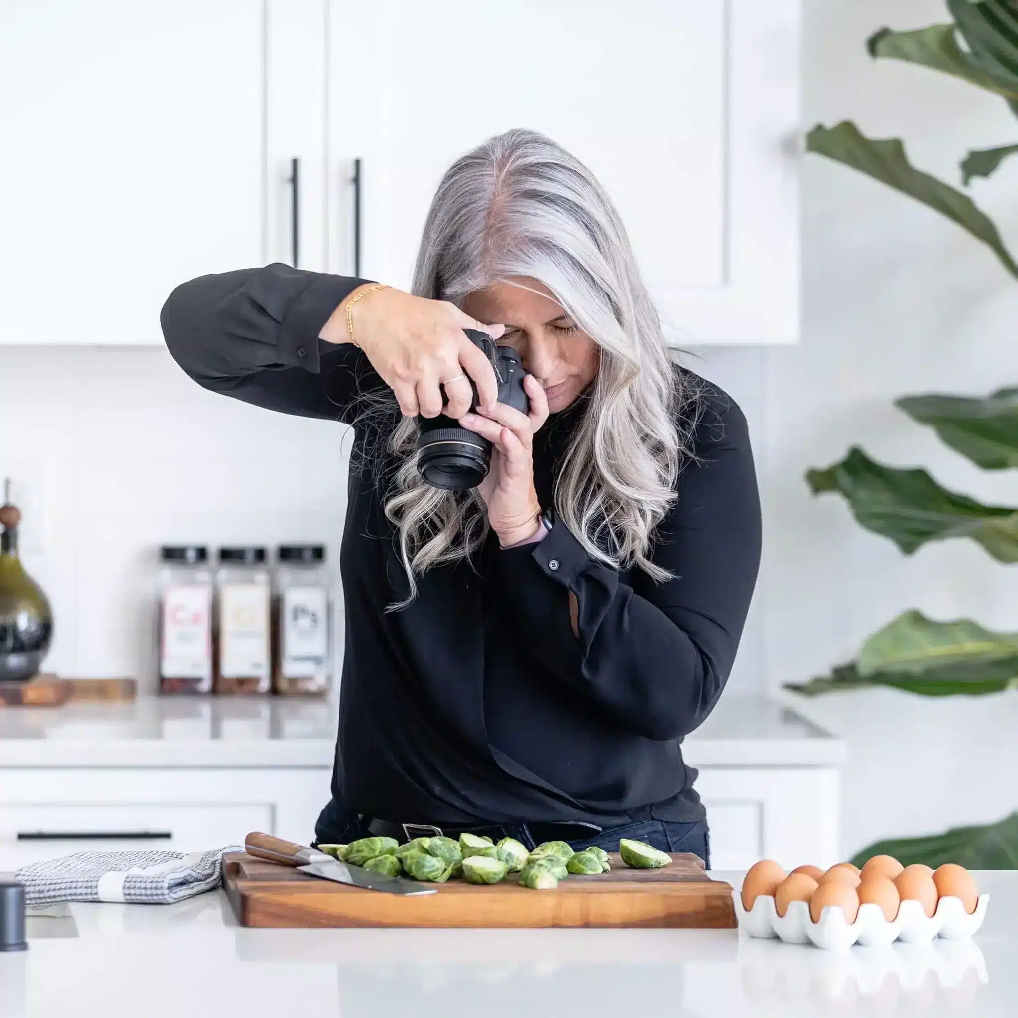A woman with long gray hair, wearing a black top, takes a photo of sliced Brussels sprouts on a wooden board in a bright kitchen, capturing her farmgirl gourmet touch. Eggs, spices, and a plant are visible in the background.