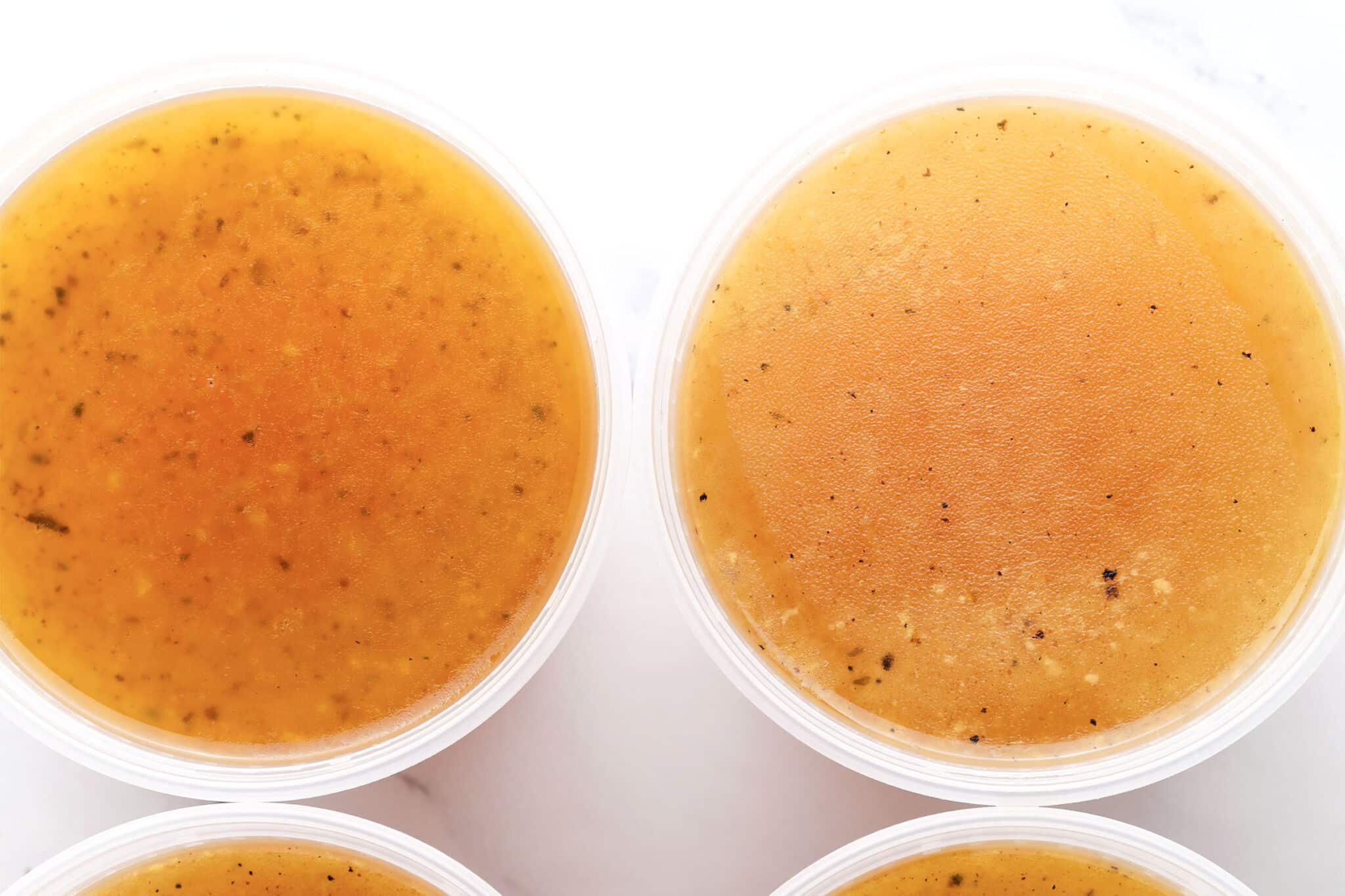 Four round plastic containers filled with golden brown broth or stock, viewed from above; the liquids have visible seasoning and slight variations in color and opacity, perfect for a basic chicken stock recipe.