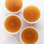 Four round, clear plastic containers filled with brown broth—made from a basic chicken stock recipe—are arranged on a white marble surface. The lids are off and placed nearby, with flecks of seasoning visible in the savory broth.