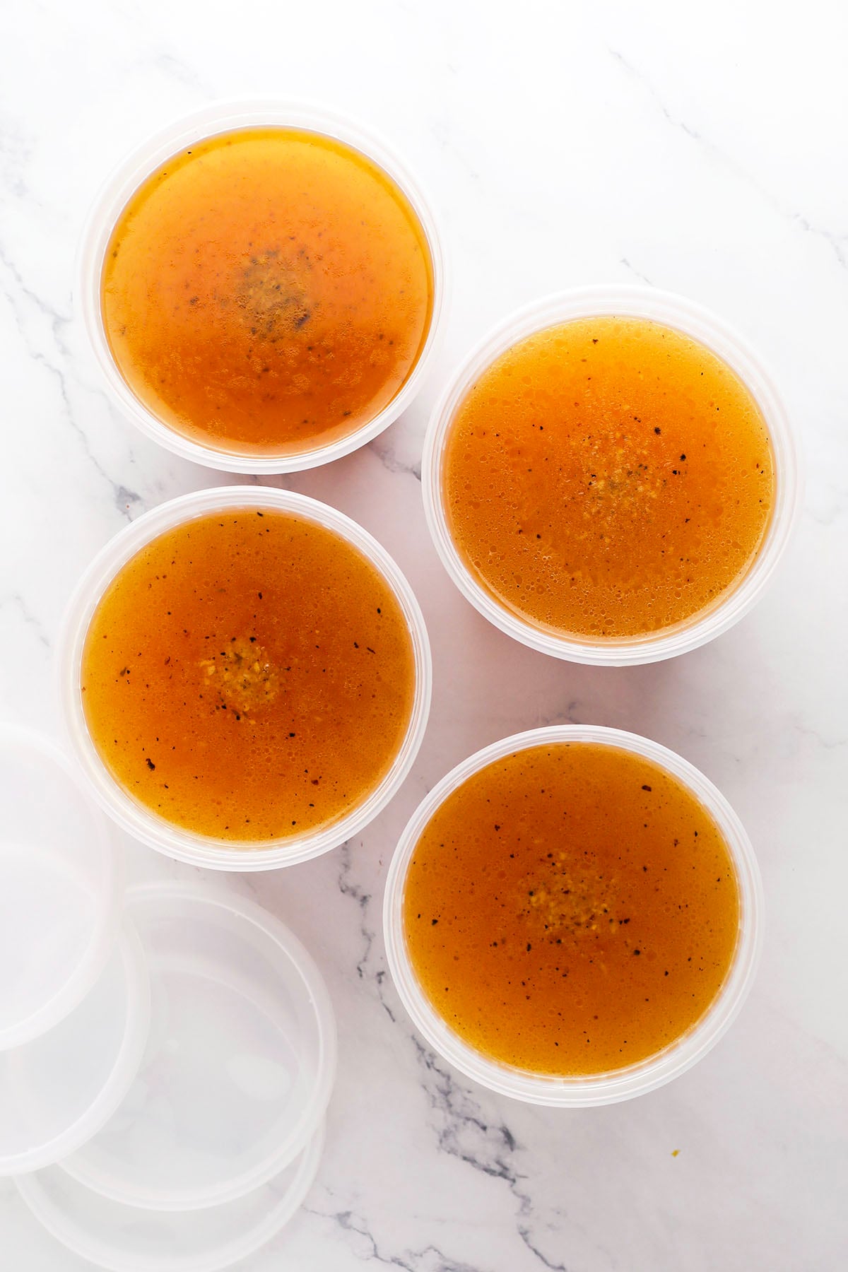 Four round, clear plastic containers filled with brown broth—made from a basic chicken stock recipe—are arranged on a white marble surface. The lids are off and placed nearby, with flecks of seasoning visible in the savory broth.