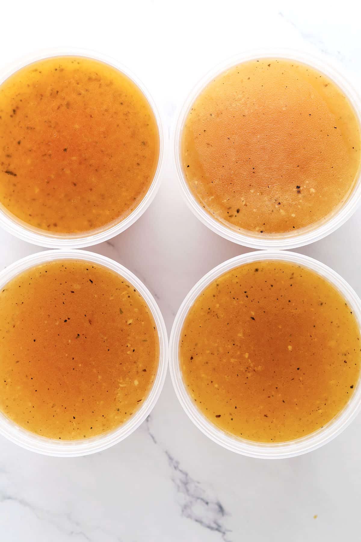 Four clear plastic containers filled with golden-brown broth, made from a basic chicken stock recipe and dotted with herbs and seasonings, arranged in a two-by-two grid on a white marble surface.