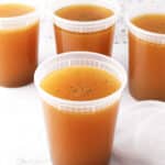 Four clear plastic containers filled with golden-brown basic chicken stock are arranged on a white surface, with one lid placed beside a container. The background features a light-colored, speckled pattern.