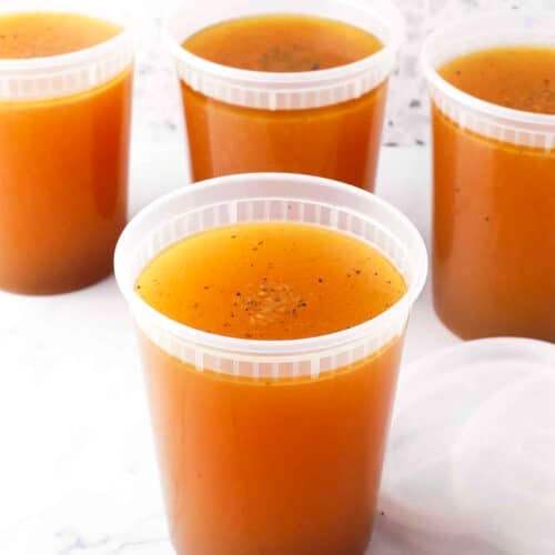 Four clear plastic containers filled with golden-brown basic chicken stock are arranged on a white surface, with one lid placed beside a container. The background features a light-colored, speckled pattern.