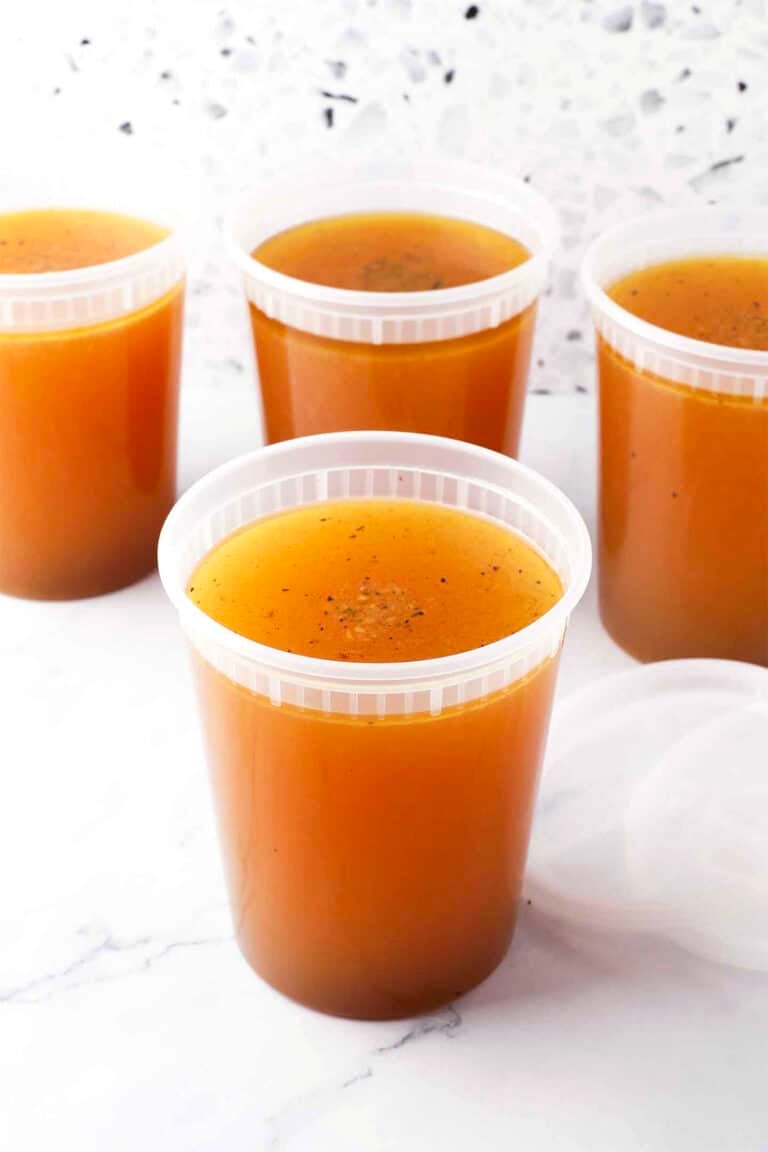 Four clear plastic containers filled with golden-brown basic chicken stock are arranged on a white surface, with one lid placed beside a container. The background features a light-colored, speckled pattern.