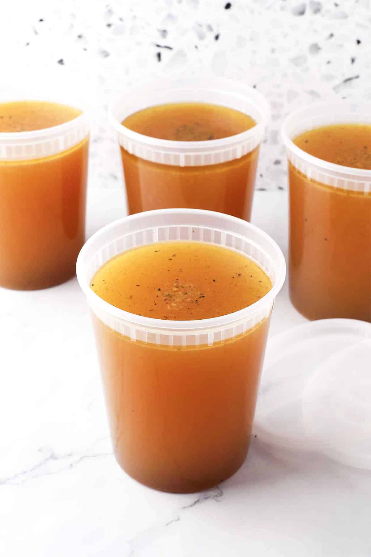 Four clear plastic containers filled with golden-brown basic chicken stock are arranged on a white surface, with one lid placed beside a container. The background features a light-colored, speckled pattern.