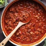 A large pot of chunky chili with beans, meat, and tomatoes sits on a dark surface, promising the best chili you’ll ever eat. A wooden spoon rests inside, ready to serve alongside bowls of chopped green onions and other tempting toppings nearby.
