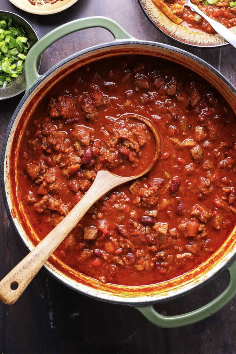 A large pot of chunky chili with beans, meat, and tomatoes sits on a dark surface, promising the best chili you’ll ever eat. A wooden spoon rests inside, ready to serve alongside bowls of chopped green onions and other tempting toppings nearby.