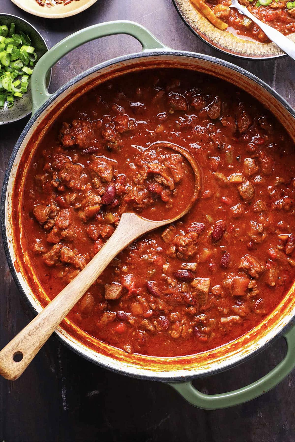 A large pot of chunky chili with beans, meat, and tomatoes sits on a dark surface, promising the best chili you’ll ever eat. A wooden spoon rests inside, ready to serve alongside bowls of chopped green onions and other tempting toppings nearby.