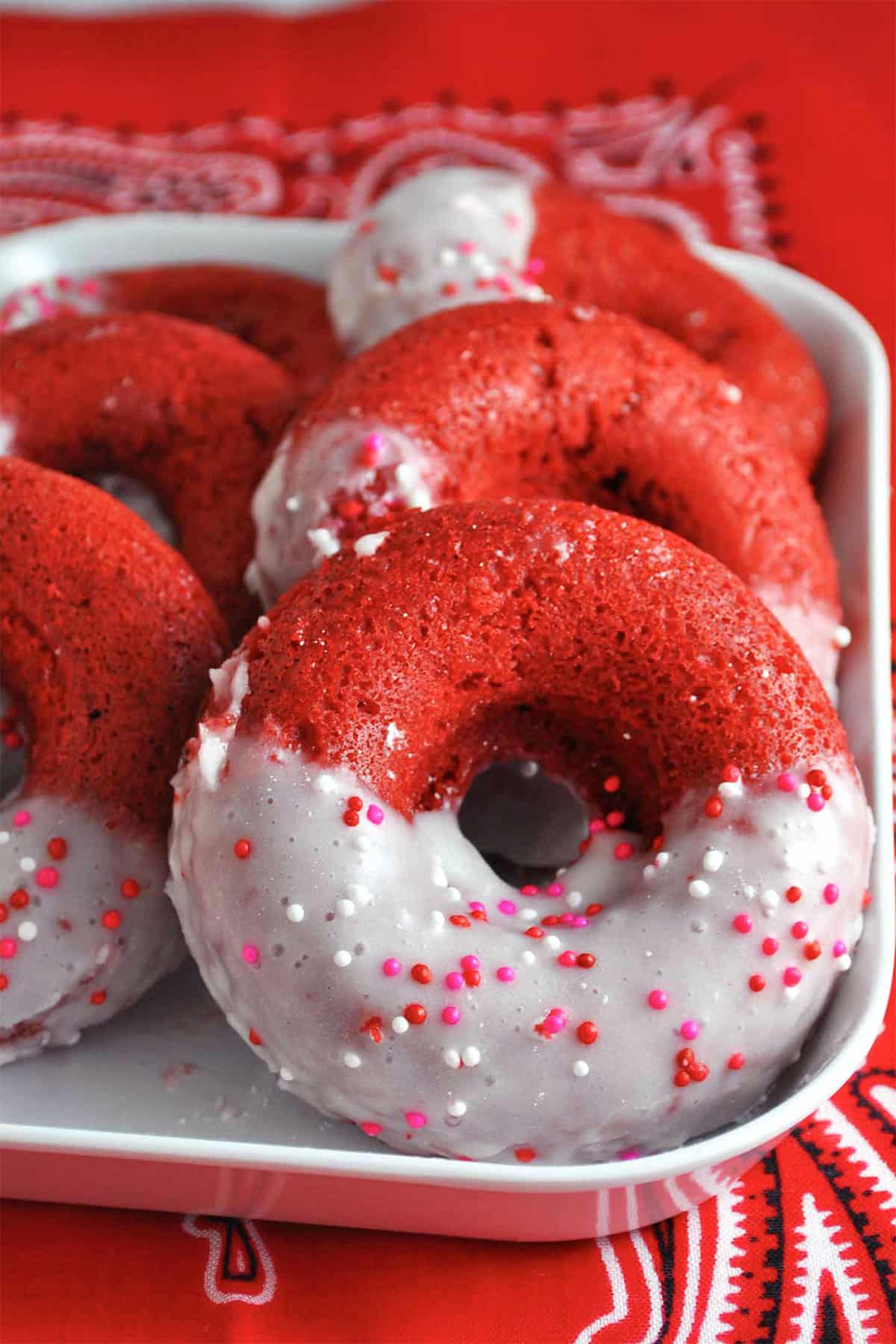A plate of baked red velvet donuts, partially dipped in white glaze and topped with pink, red, and white sprinkles, set on a red patterned cloth.