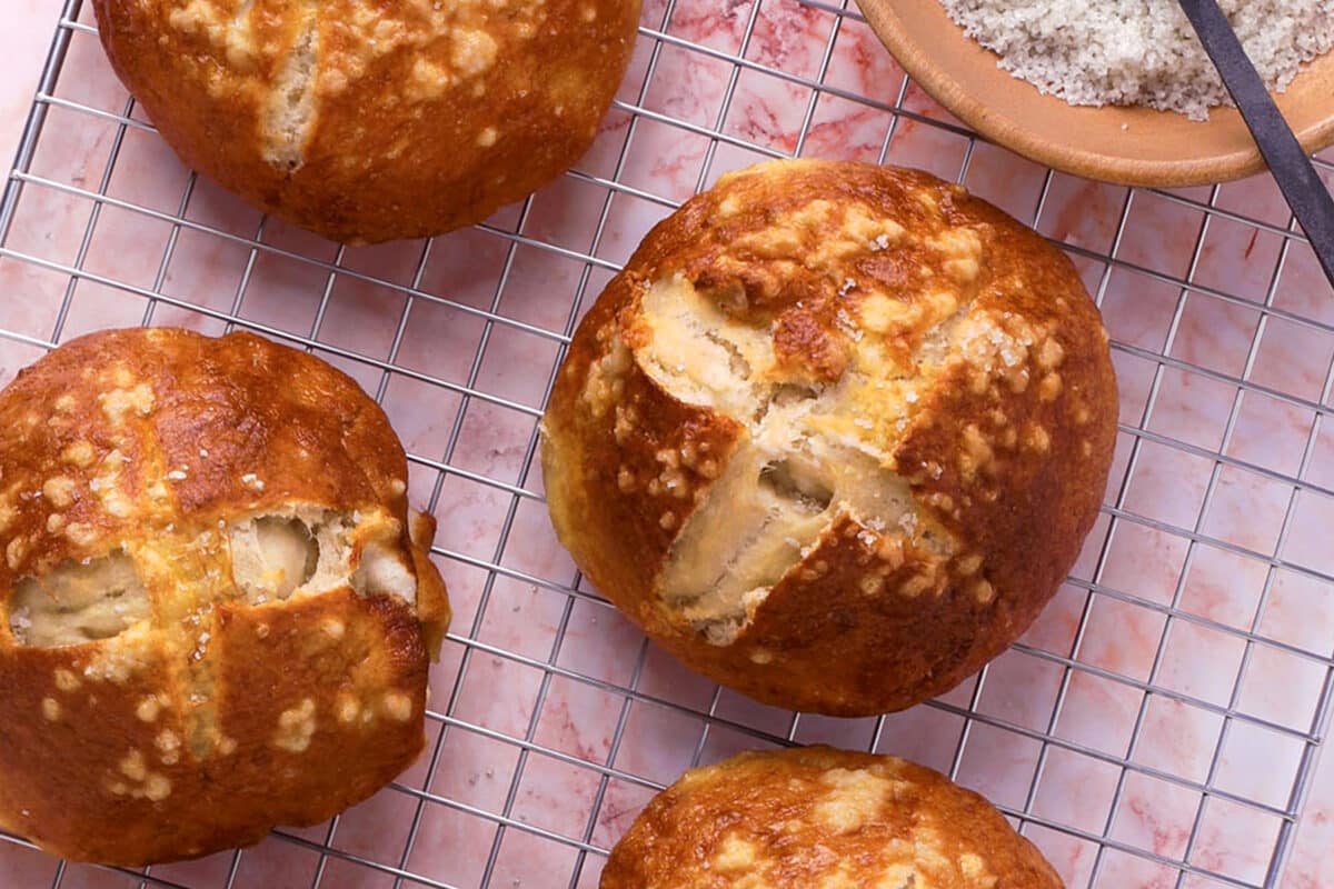 Four golden-brown pretzel burger buns with cross-shaped cuts on top rest on a cooling rack, with a bowl of coarse salt nearby.