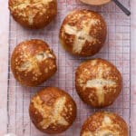 Six golden brown pretzel burger buns with salt on top, cooling on a wire rack. A small bowl of coarse salt with a black spoon is placed at the top right of the image. A white cloth is in the lower left corner.