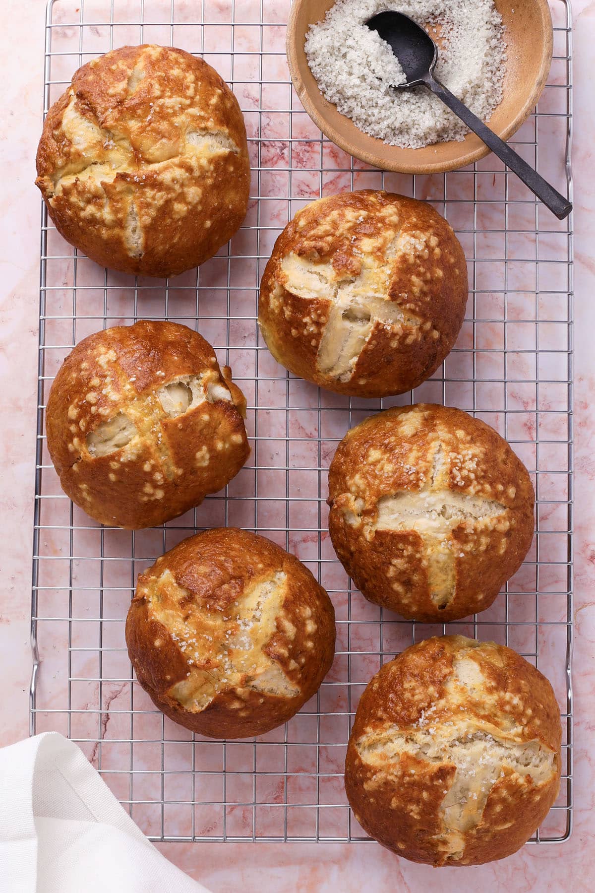 Six golden brown pretzel burger buns with salt on top, cooling on a wire rack. A small bowl of coarse salt with a black spoon is placed at the top right of the image. A white cloth is in the lower left corner.