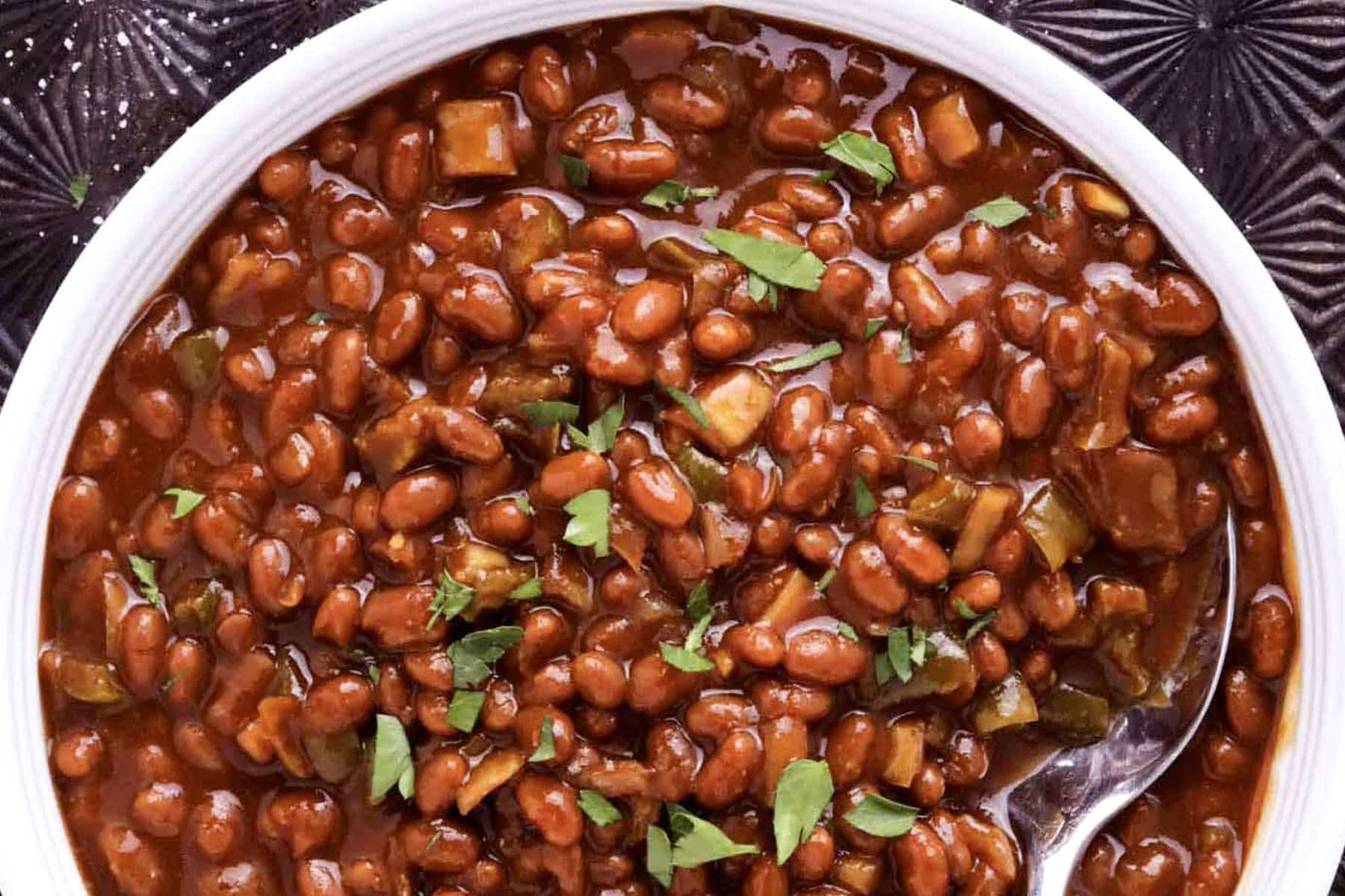 A close-up of a bowl of easy slow cooker baked beans in a rich sauce, garnished with chopped fresh herbs, with a spoon partially visible on the right side.