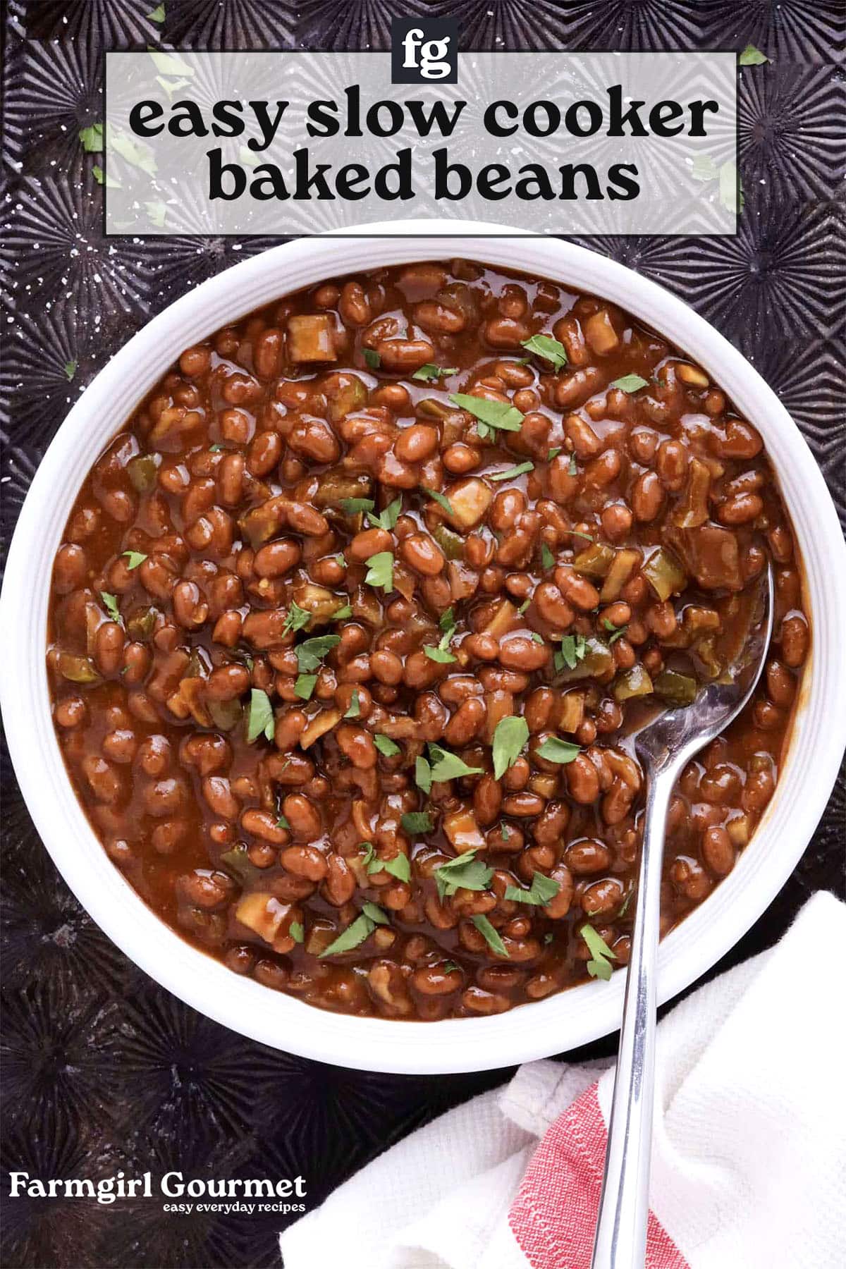 A bowl of easy slow cooker baked beans, garnished with chopped herbs, sits on a dark textured surface with a spoon resting inside. A red-striped towel is nearby, and text on the image reads: easy slow cooker baked beans.
