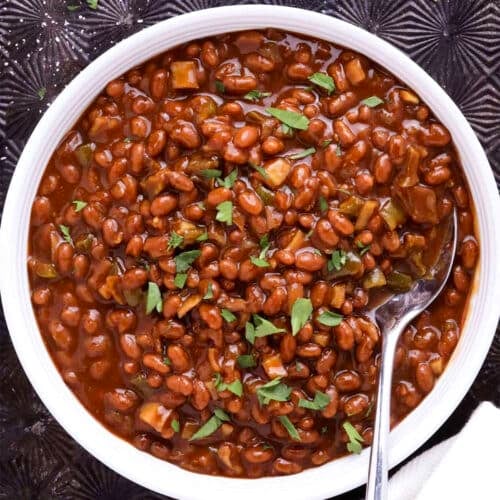 A white bowl filled with easy slow cooker baked beans in a thick, brown sauce, garnished with chopped herbs. A spoon rests inside the bowl, and a white and red towel is beside it on a dark textured surface.