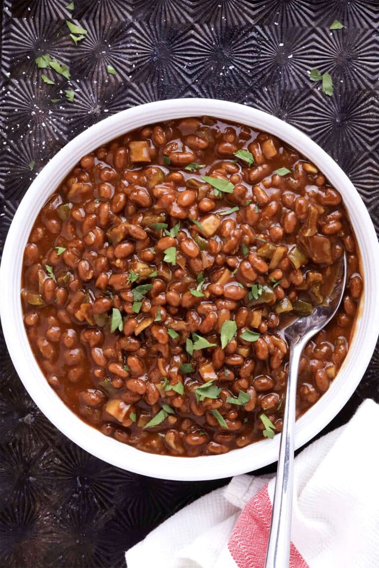 A white bowl filled with easy slow cooker baked beans in a thick, brown sauce, garnished with chopped herbs. A spoon rests inside the bowl, and a white and red towel is beside it on a dark textured surface.