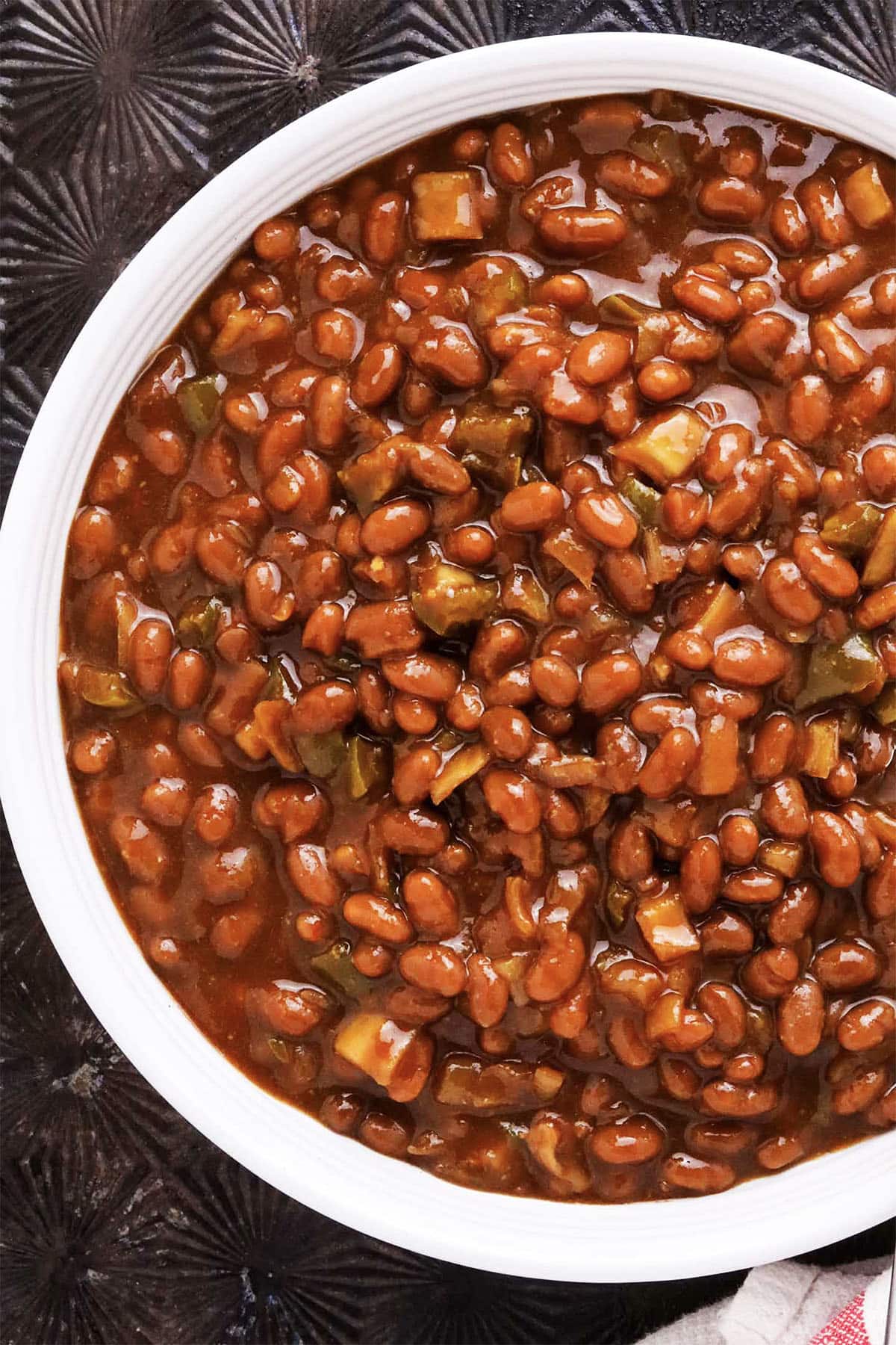 A white bowl filled with easy slow cooker baked beans in a rich, brown sauce, mixed with small pieces of green bell pepper and onion, set on a dark textured surface.