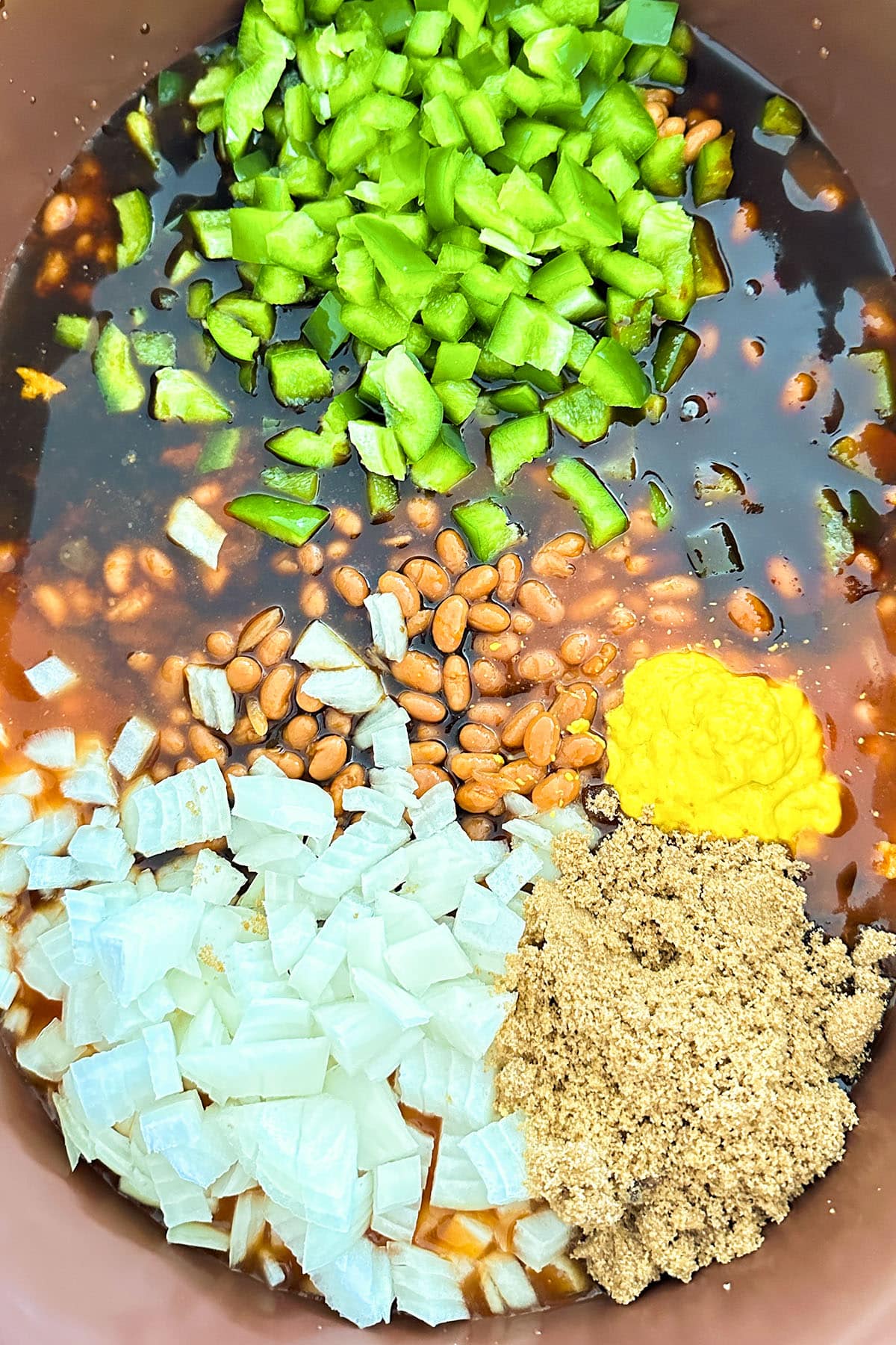 Chopped green bell pepper, diced onions, pinto beans, yellow mustard, brown sugar, and a dark sauce are arranged in sections in a mixing bowl—perfect for making easy slow cooker baked beans.
