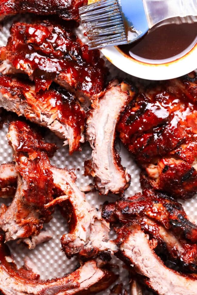 Barbecued ribs with a glossy, caramelized sauce—truly the best BBQ'd ribs—are arranged on a tray. A basting brush and a small bowl of barbecue sauce sit in the corner. The ribs look juicy and tender.