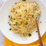 A white plate of lemon angel hair pasta garnished with chopped parsley, lemon zest, and cracked black pepper sits on a yellow napkin, with a fork resting on the side of the plate.
