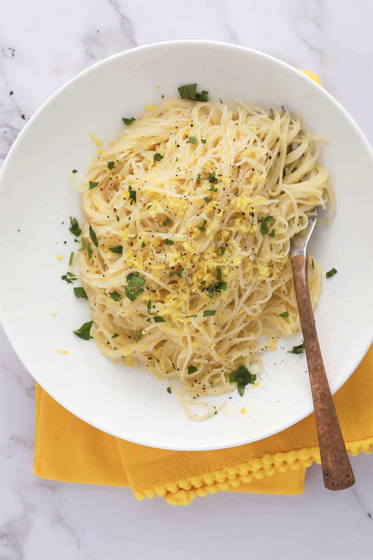 A white plate of lemon angel hair pasta garnished with chopped parsley, lemon zest, and cracked black pepper sits on a yellow napkin, with a fork resting on the side of the plate.