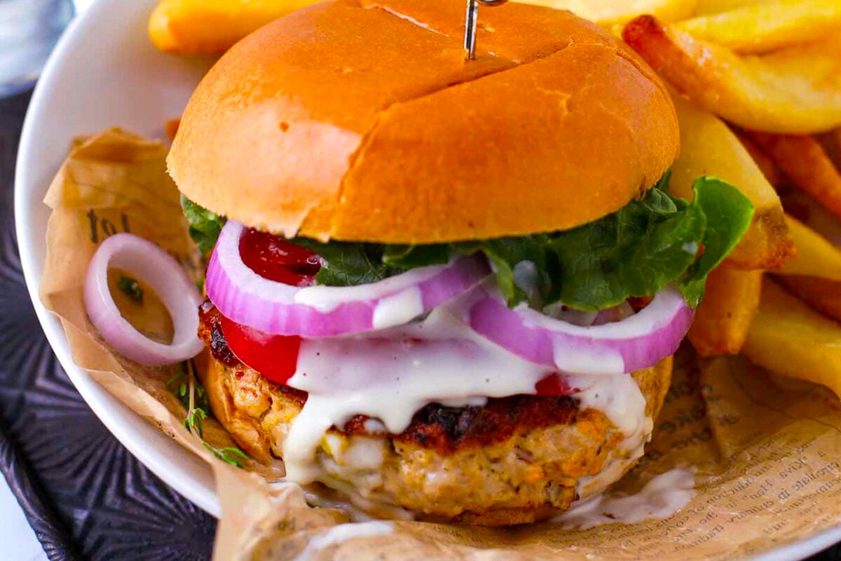 A close-up of buffalo chicken burgers with lettuce, tomato, red onion, and white sauce in a bun, served with thick-cut fries on the side. The burger is placed on a paper-lined plate.