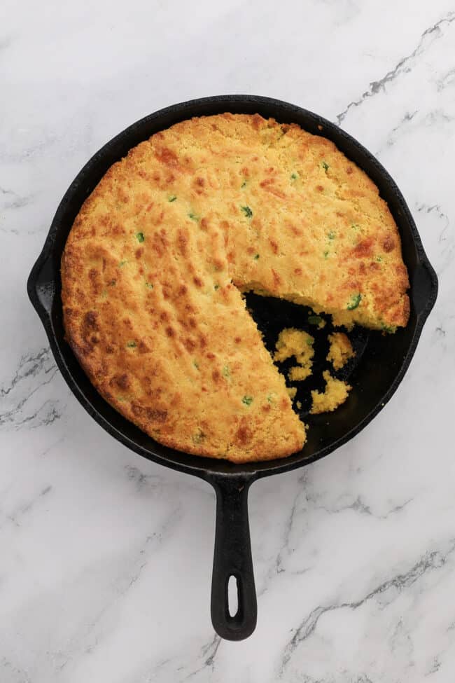 A round skillet of golden brown jalapeno cheddar cornbread with green flecks, missing one wedge-shaped slice, sits on a white marble surface. Crumbs are visible in the empty section of the pan.