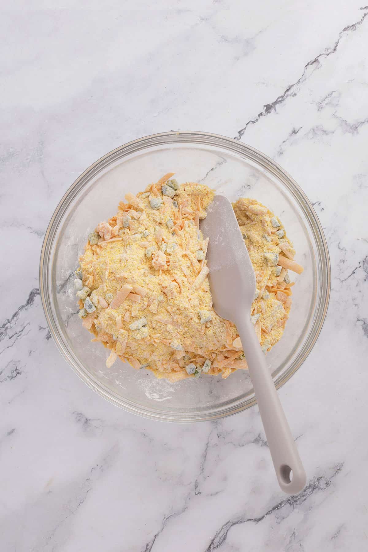 A glass mixing bowl containing yellow, crumbly jalapeno cheddar cornbread dough with visible chunks and a gray spatula on a light marble surface.