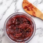 A small glass jar filled with chunky red strawberry jam sits on a marble surface, accompanied by a wooden spoon with jam residue and a hint of rosemary fig jam nearby.