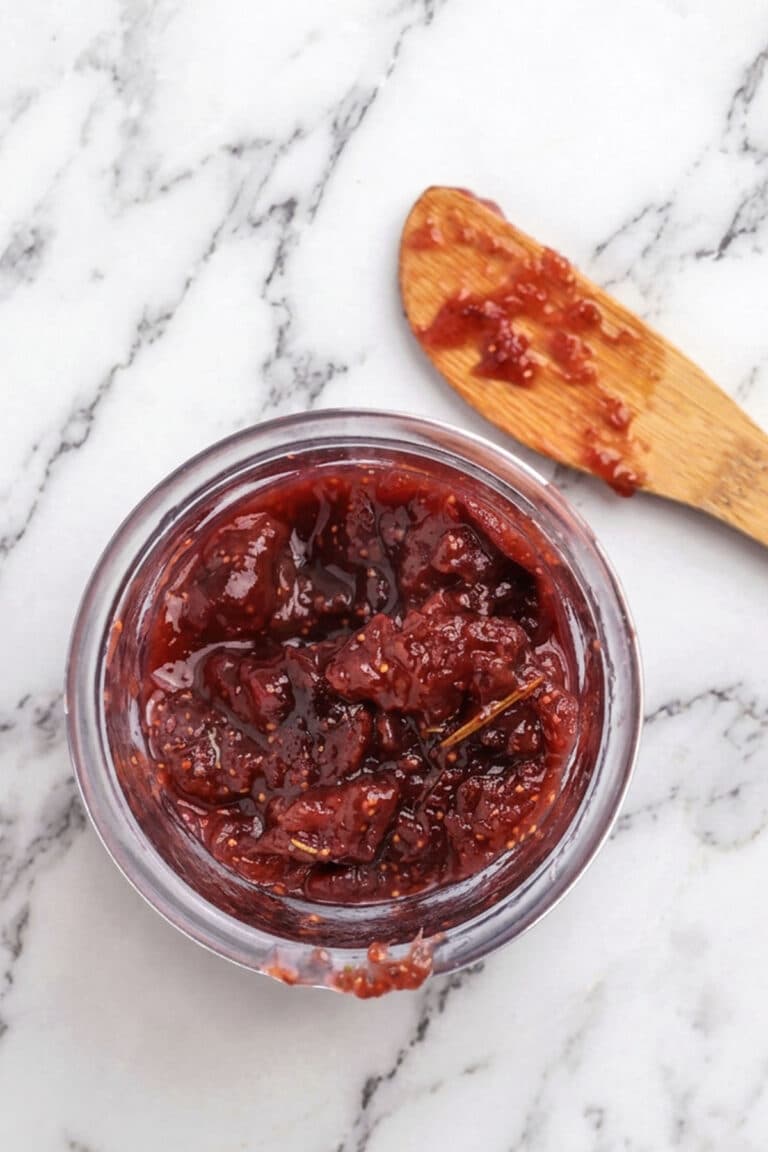 A small glass jar filled with chunky red strawberry jam sits on a marble surface, accompanied by a wooden spoon with jam residue and a hint of rosemary fig jam nearby.