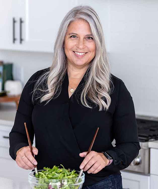 Smiling woman with long gray hair stands in a kitchen, wearing a black blouse, holding wooden salad tongs and a bowl of salad with greens. Modern white kitchen background with stove and cabinets.