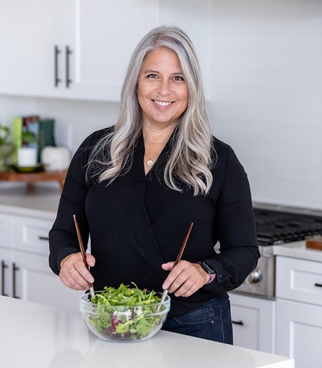 A woman with long gray hair, wearing a black shirt, stands in a modern kitchen smiling while tossing a salad with wooden utensils in a glass bowl on a white countertop.