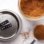 A jar of chai spice blend with its lid labeled CHAI SPICE, some of the spice spilled, and a tablespoon filled with chai spice on a white marble surface.