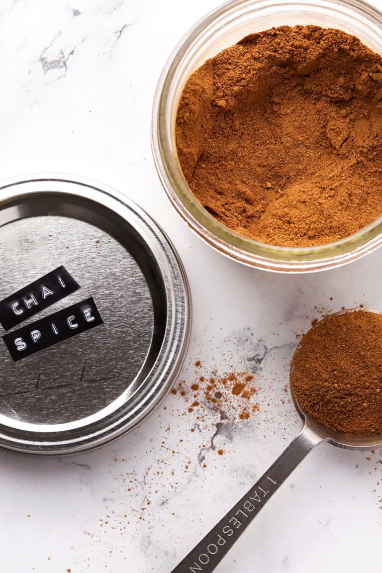 A jar of chai spice blend with its lid labeled CHAI SPICE, some of the spice spilled, and a tablespoon filled with chai spice on a white marble surface.