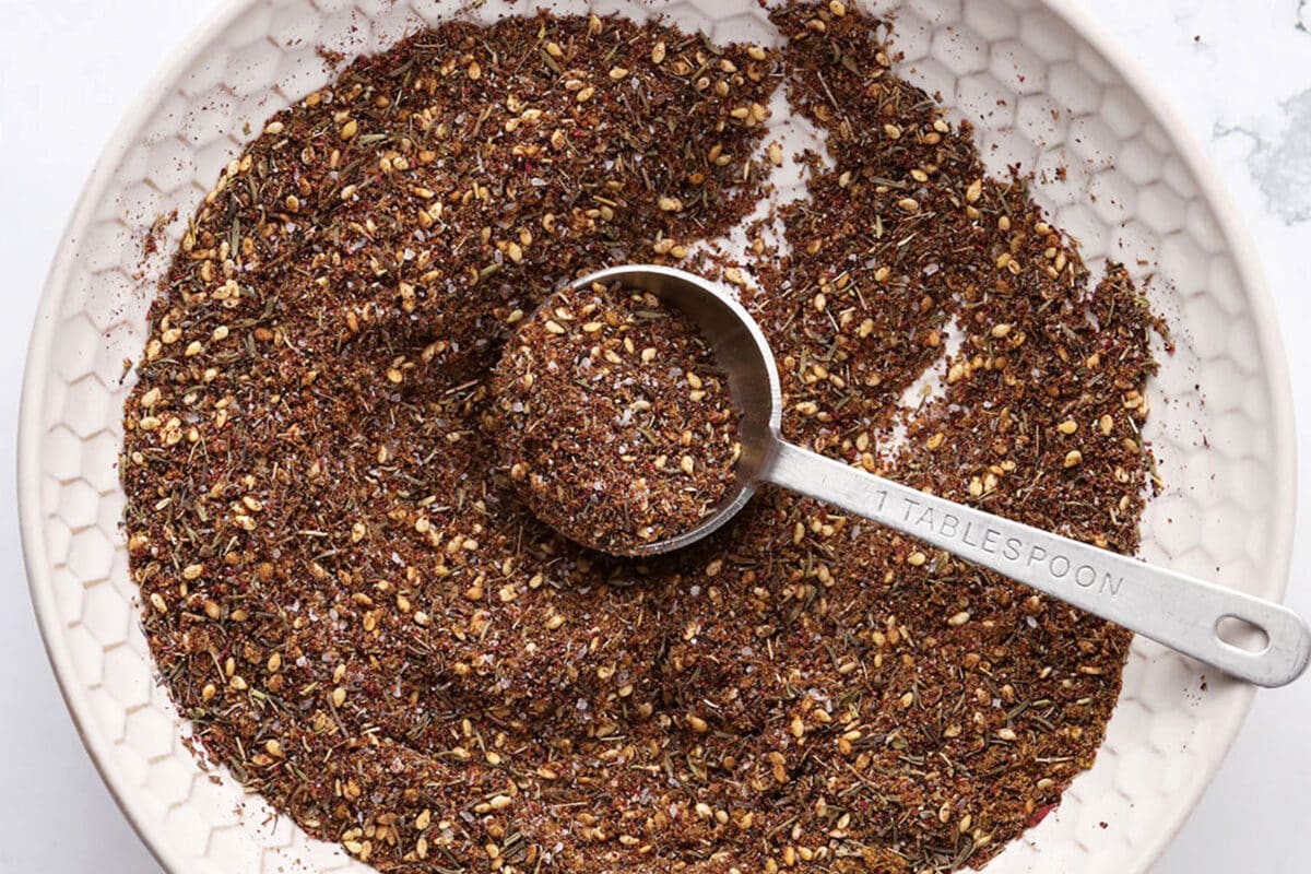 A white bowl holds a coarse, brown za'atar spice blend with visible seeds and herbs. A metal tablespoon labeled 1 TABLESPOON rests in the bowl, scooping up the aromatic mixture.