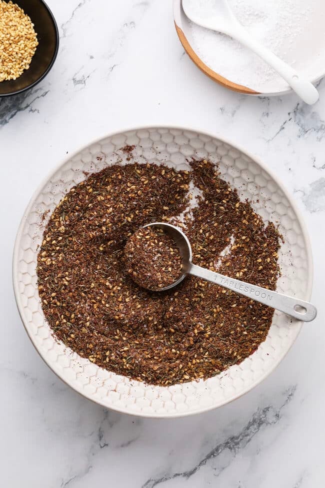 A bowl filled with a coarse, brown za'atar spice blend sits on a white surface. A metal tablespoon rests inside, holding some of the mix. Small bowls with sesame seeds and salt are nearby—perfect for learning how to make za’atar at home.