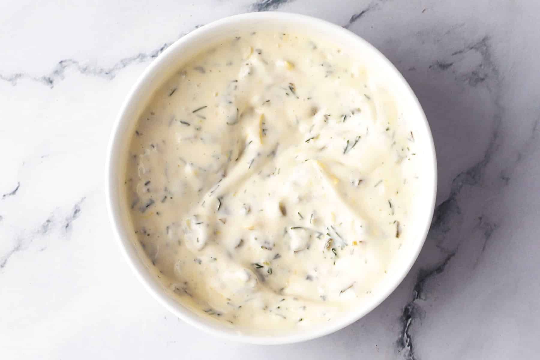 A white bowl filled with creamy, herb-flecked basic tartar sauce sits on a white marble surface.