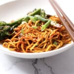 A plate of stir-fried noodles topped with sesame seeds and sliced green onions, served with broccolini and chopsticks on a white marble surface.