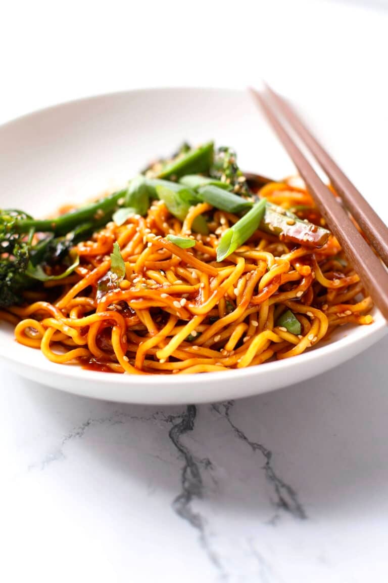 A plate of stir-fried noodles topped with sesame seeds and sliced green onions, served with broccolini and chopsticks on a white marble surface.