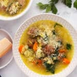 Overhead view of a bowl of soup with meatballs, carrots, leafy greens, grated cheese, and a light broth, garnished with parsley. A block of cheese and fresh parsley are nearby on a marble surface.