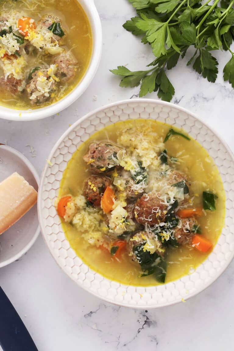 Overhead view of a bowl of soup with meatballs, carrots, leafy greens, grated cheese, and a light broth, garnished with parsley. A block of cheese and fresh parsley are nearby on a marble surface.