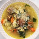 A bowl of Italian wedding soup with meatballs, carrots, spinach, and rice in a clear broth, garnished with grated cheese. The comforting Italian wedding soup sits on a white surface with parsley nearby.