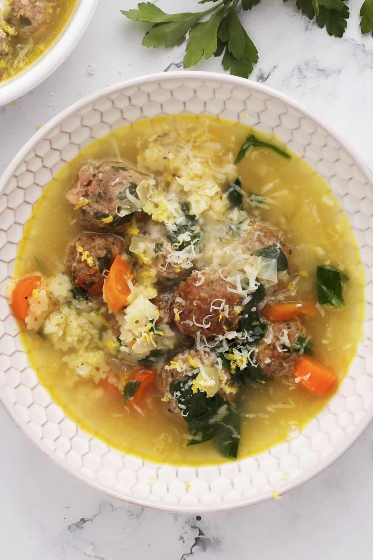 A bowl of soup with meatballs, carrots, spinach, and orzo, garnished with grated cheese and lemon zest. Fresh parsley is visible in the background.