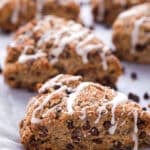 A close-up of chocolate chip scones drizzled with white icing, arranged on a baking sheet lined with parchment paper. More scones are visible in the background.