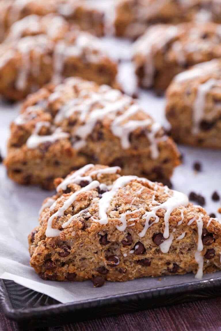 A close-up of chocolate chip scones drizzled with white icing, arranged on a baking sheet lined with parchment paper. More scones are visible in the background.