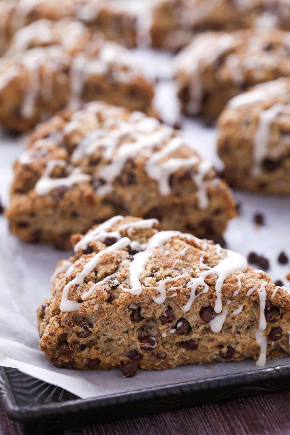 A close-up of chocolate chip scones drizzled with white icing, arranged on a baking sheet lined with parchment paper. More scones are visible in the background.