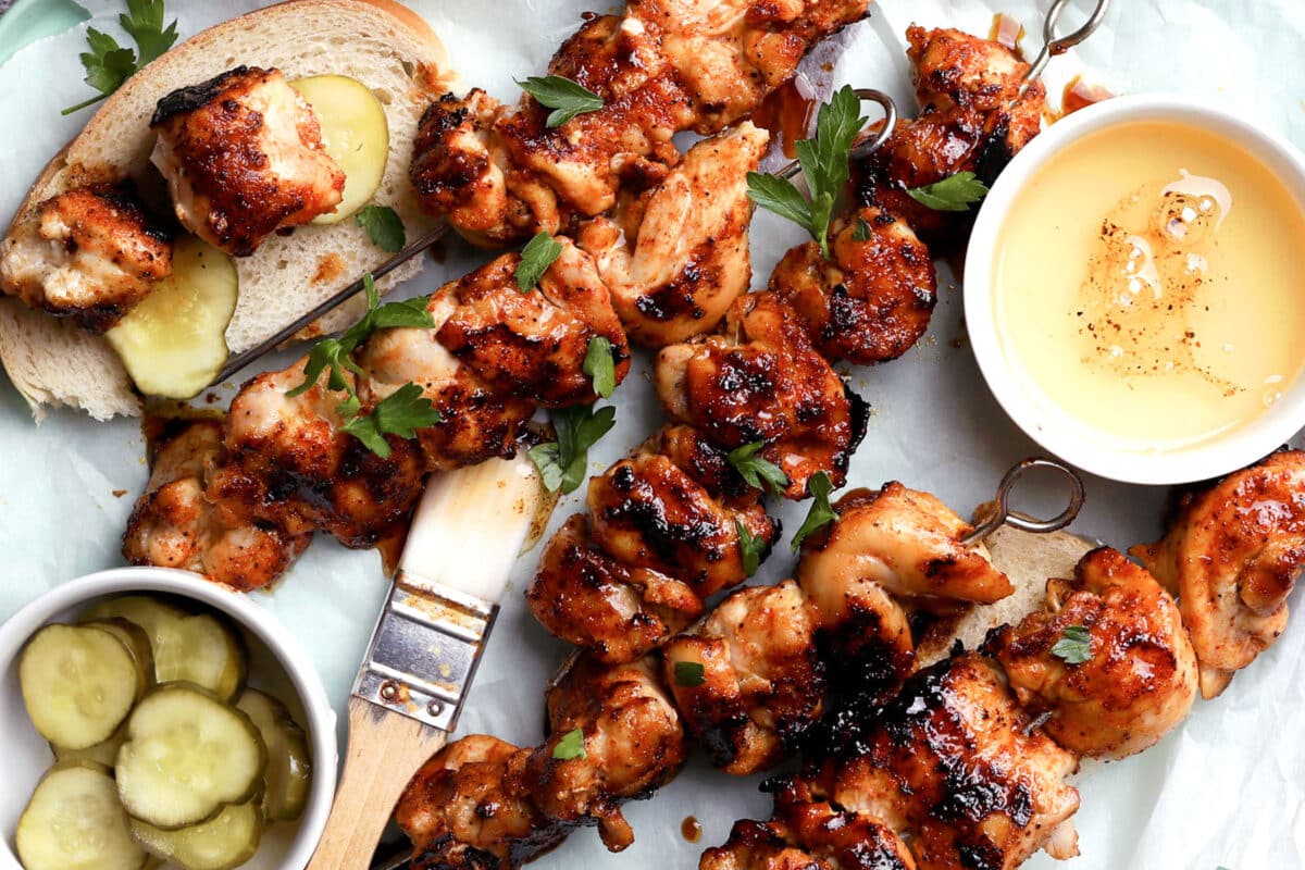 Grilled Nashville hot chicken skewers with a glaze are served on parchment with sliced bread, pickles, fresh parsley, a bowl of honey mustard dip, and a basting brush.