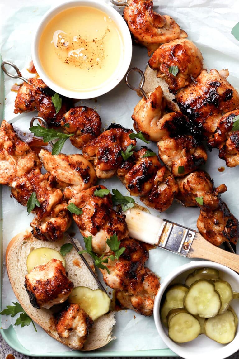 Grilled Nashville hot chicken skewers with a glazed finish are served on slices of bread with pickles, garnished with parsley. A brush, a bowl of honey, and a small bowl of pickles are also on the tray.
